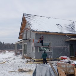 Oleksandr Vasiliev DACHOLEK - Dom w trakcie ocieplania, widoczne rusztowania i pracownicy montujący izolację. Dach pokryty śniegiem, zimowy krajobraz w tle.