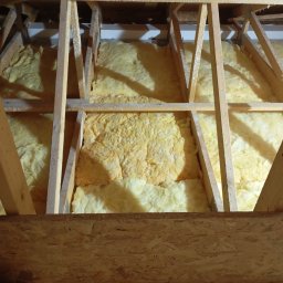 Oleksandr Vasiliev DACHOLEK - Ocieplenie poddasza wełną mineralną, widoczne drewniane elementy konstrukcyjne dachu. Fragment izolacji z bliska, ciepłe barwy oświetlenia.