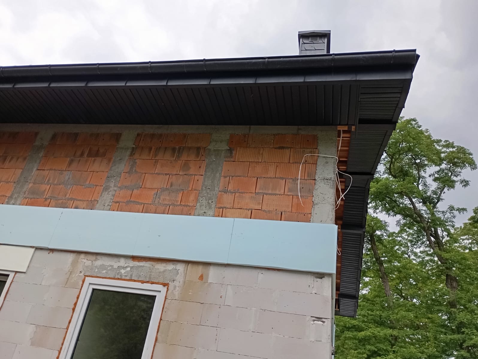 Fragment nowo wybudowanego domu z widocznym murem z czerwonej cegły, ociepleniem ze styropianu oraz zamontowanym ciemnym orynnowaniem i podbitką dachową, na tle pochmurnego nieba i drzew.