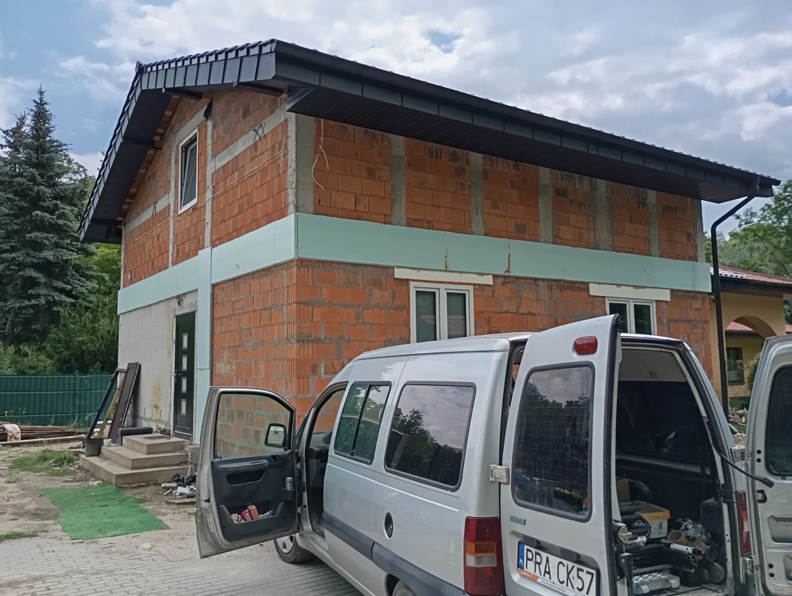 Surowy budynek w trakcie budowy z nowym, ciemnym dachem; widoczny samochód dostawczy z otwartymi drzwiami i narzędziami w środku.