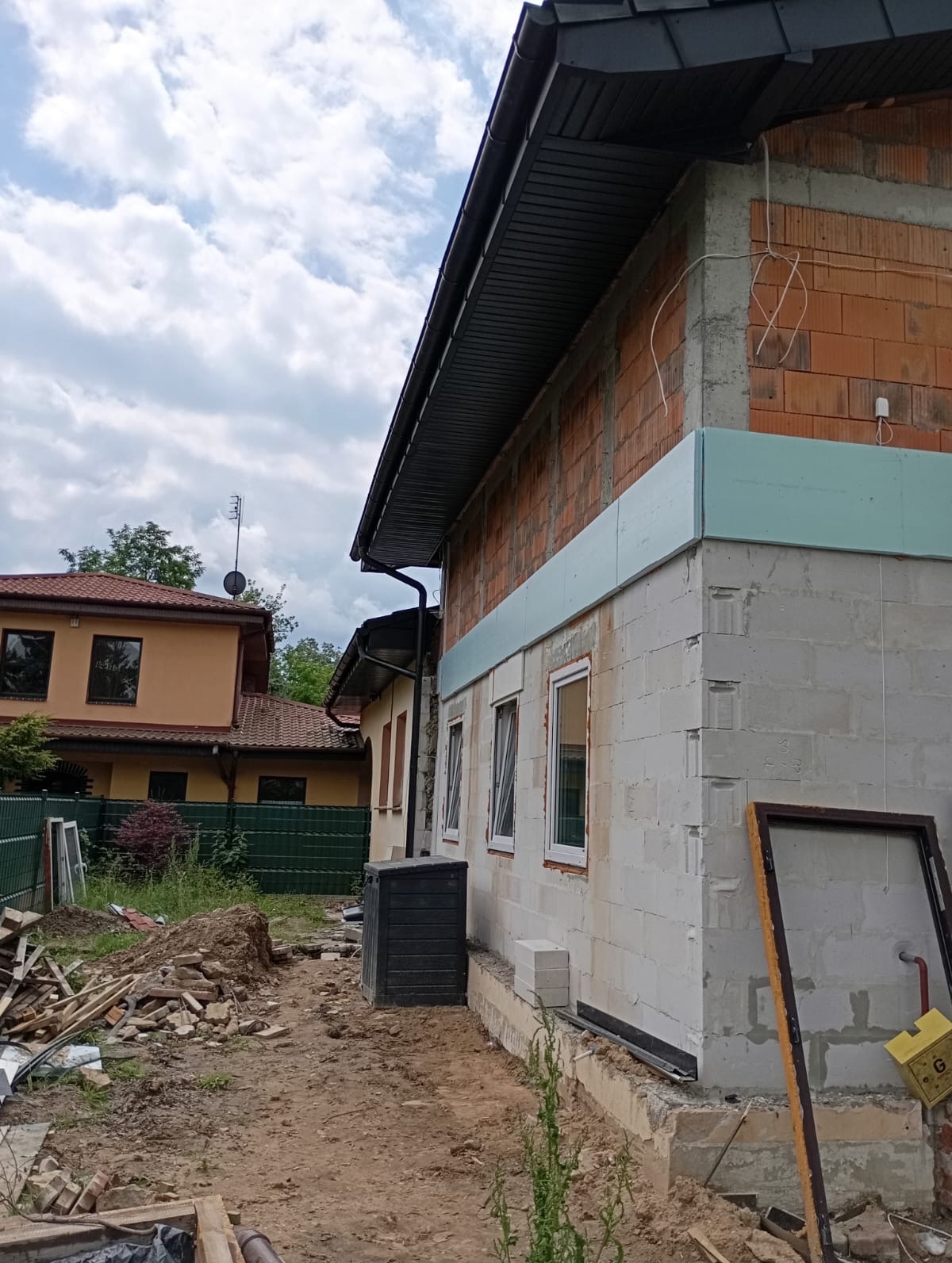 Budynek w trakcie ocieplania: widoczne bloczki, styropian, okna, rynna, oraz fragment dachu z ciemną dachówką, z elementami rozgardiaszu budowlanego w otoczeniu.