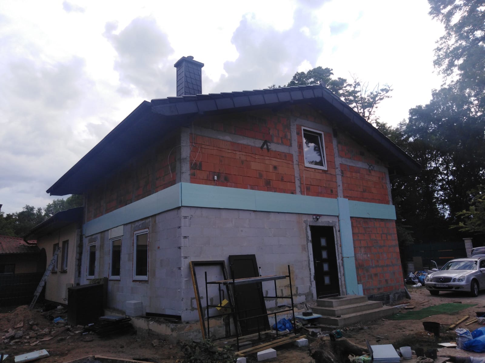 Dom w trakcie budowy: widoczne mury z czerwonej cegły, ocieplenie styropianem, czarny dach z kominem oraz teren budowy z narzędziami i materiałami.