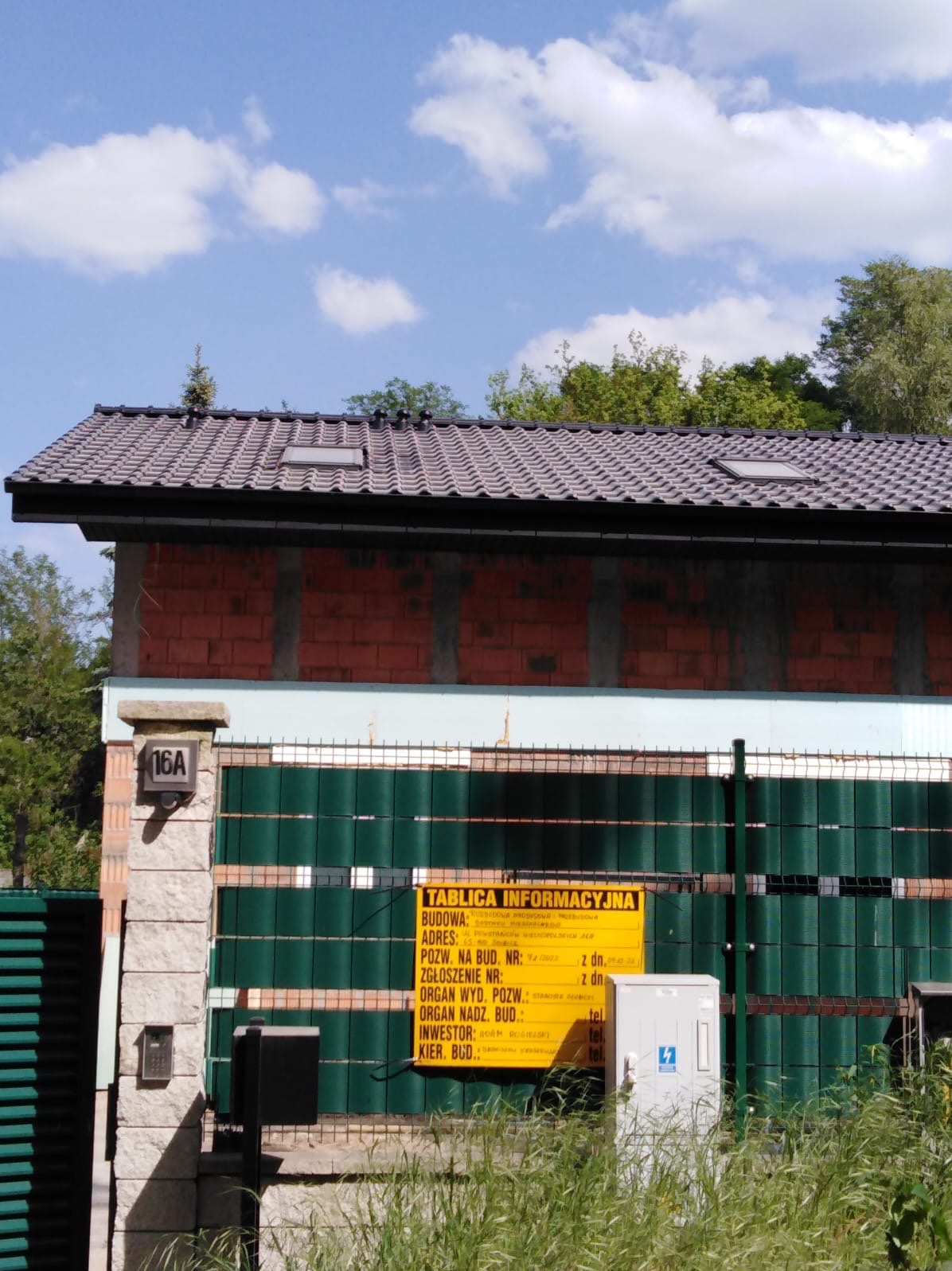 Budynek w trakcie budowy z widocznym fragmentem dachu z dachówkami, oknami dachowymi, murem z czerwonej cegły, zielonym ogrodzeniem i tablicą informacyjną budowy.
