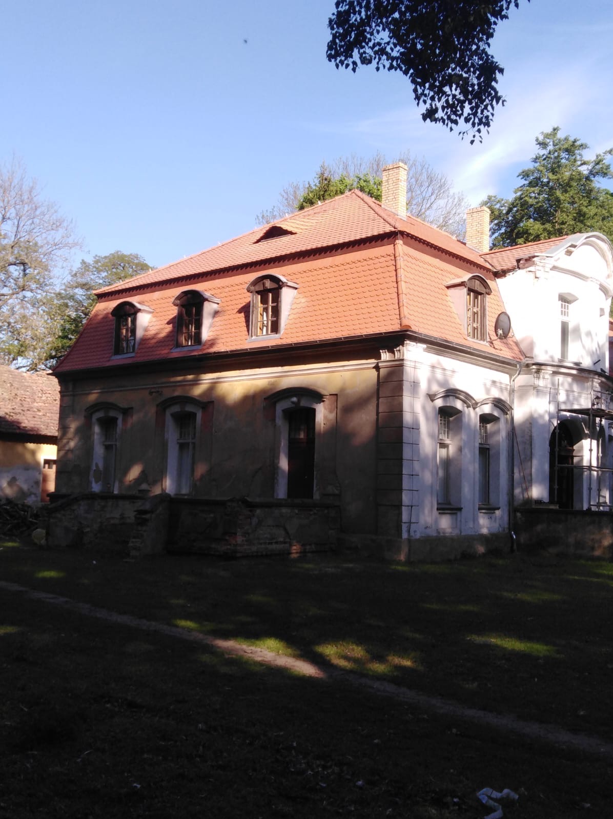 Dwukondygnacyjny, częściowo odnowiony budynek z czerwonym dachem z dachówki, widoczne okna dachowe i kominy, z fragmentami elewacji w trakcie renowacji.