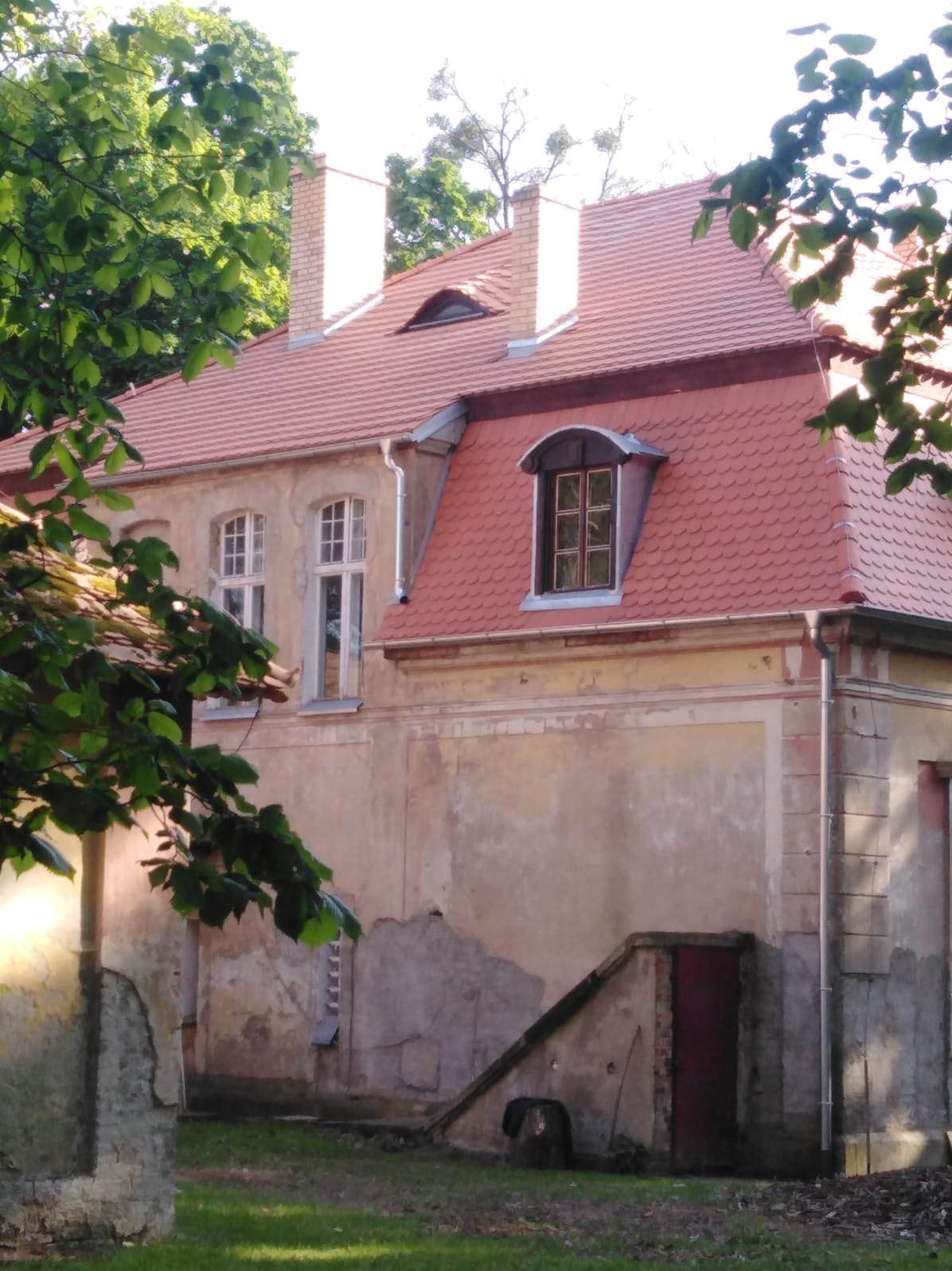 Fragment budynku z czerwonym dachem pokrytym dachówką karpiówką, widoczne okna dachowe typu wole oko i lukarna, kominy z cegły oraz rynny.