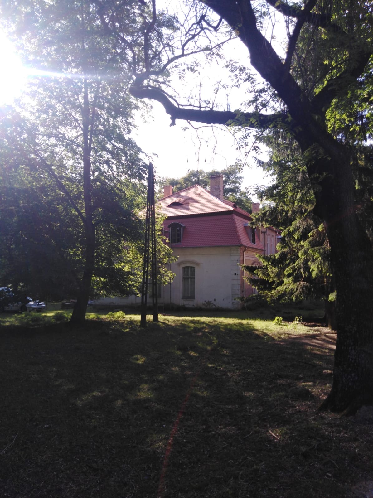 Dworek z czerwonym dachem w otoczeniu drzew, widok z trawnika w słoneczny dzień.