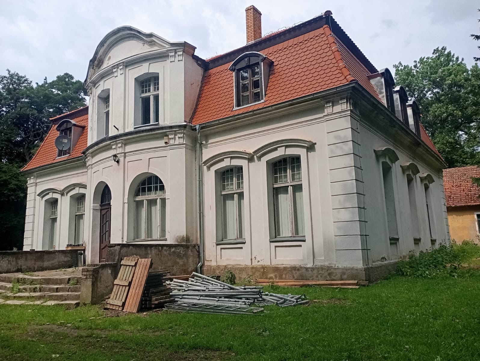 Biały, zabytkowy budynek z czerwoną dachówką, widoczne rusztowania i deski przygotowane do prac remontowych.