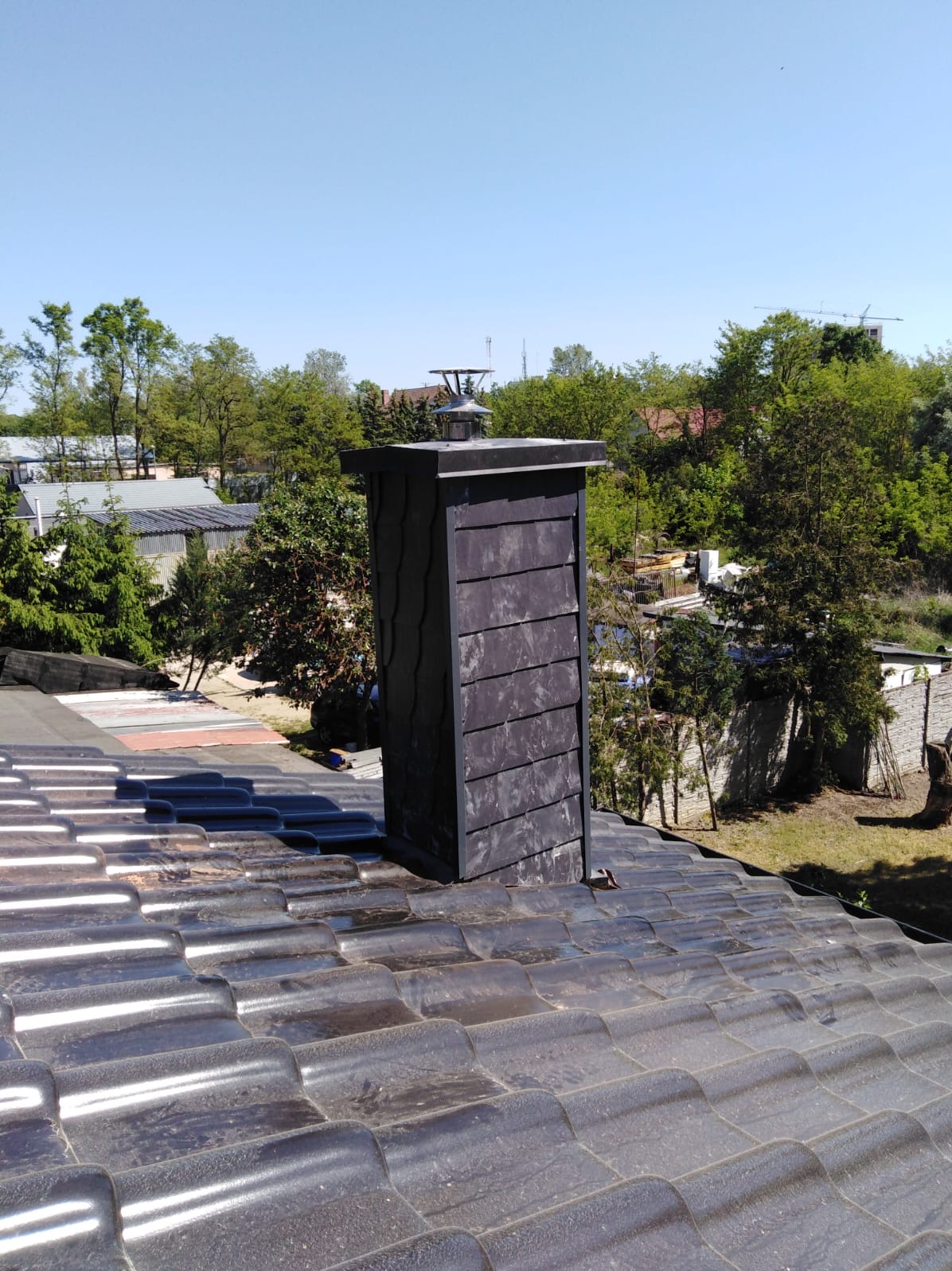 Ciemny, nowy komin z metalową czapą, wystający z błyszczącego, brązowego dachu ceramicznego w słoneczny dzień.