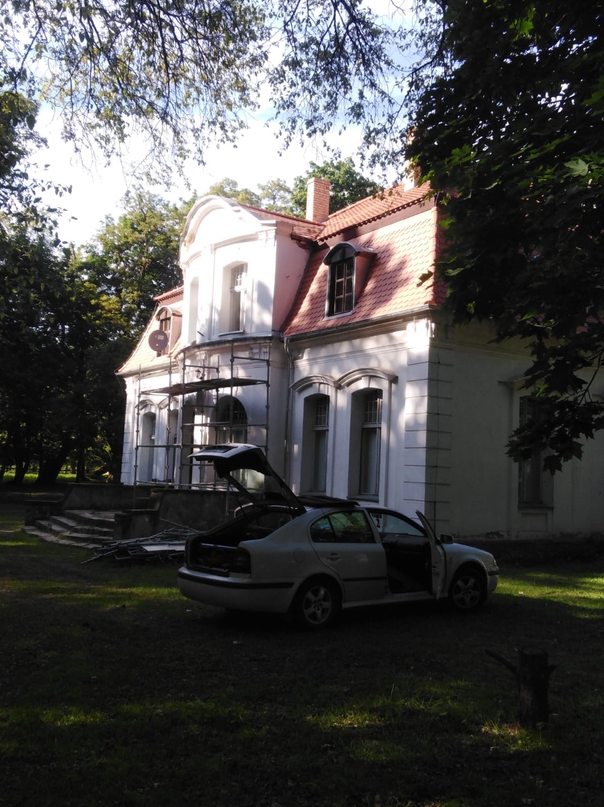 Renowacja dachu w zabytkowym domu: widoczne rusztowania, częściowo odnowiona dachówka i samochód z otwartym bagażnikiem na trawniku.