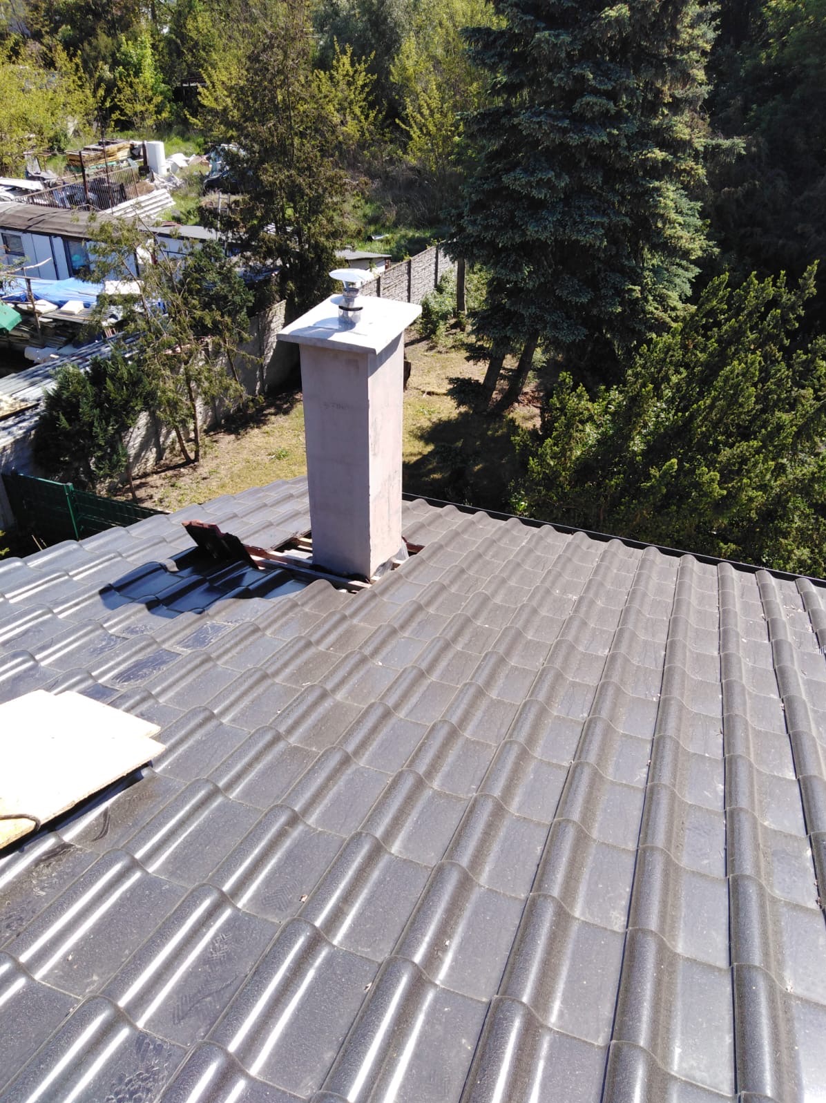 Czarny dach z dachówki ceramicznej, z widocznym kominem i niewielkimi uszkodzeniami, na tle zieleni drzew.