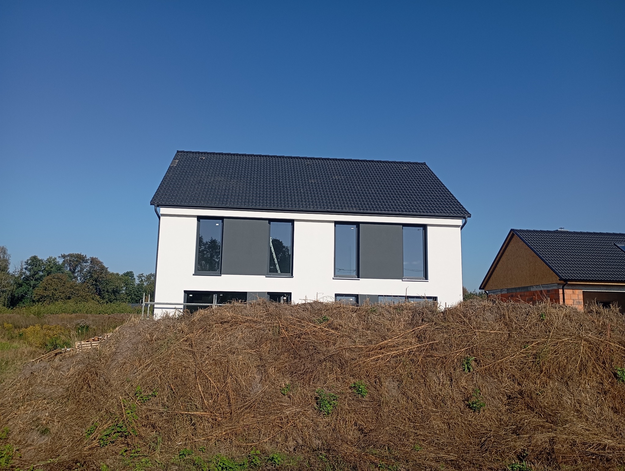 Nowoczesny dom jednorodzinny z czarną dachówką, widoczny zza kopca ziemi i uschniętych traw, z drugim budynkiem w tle w stanie surowym.