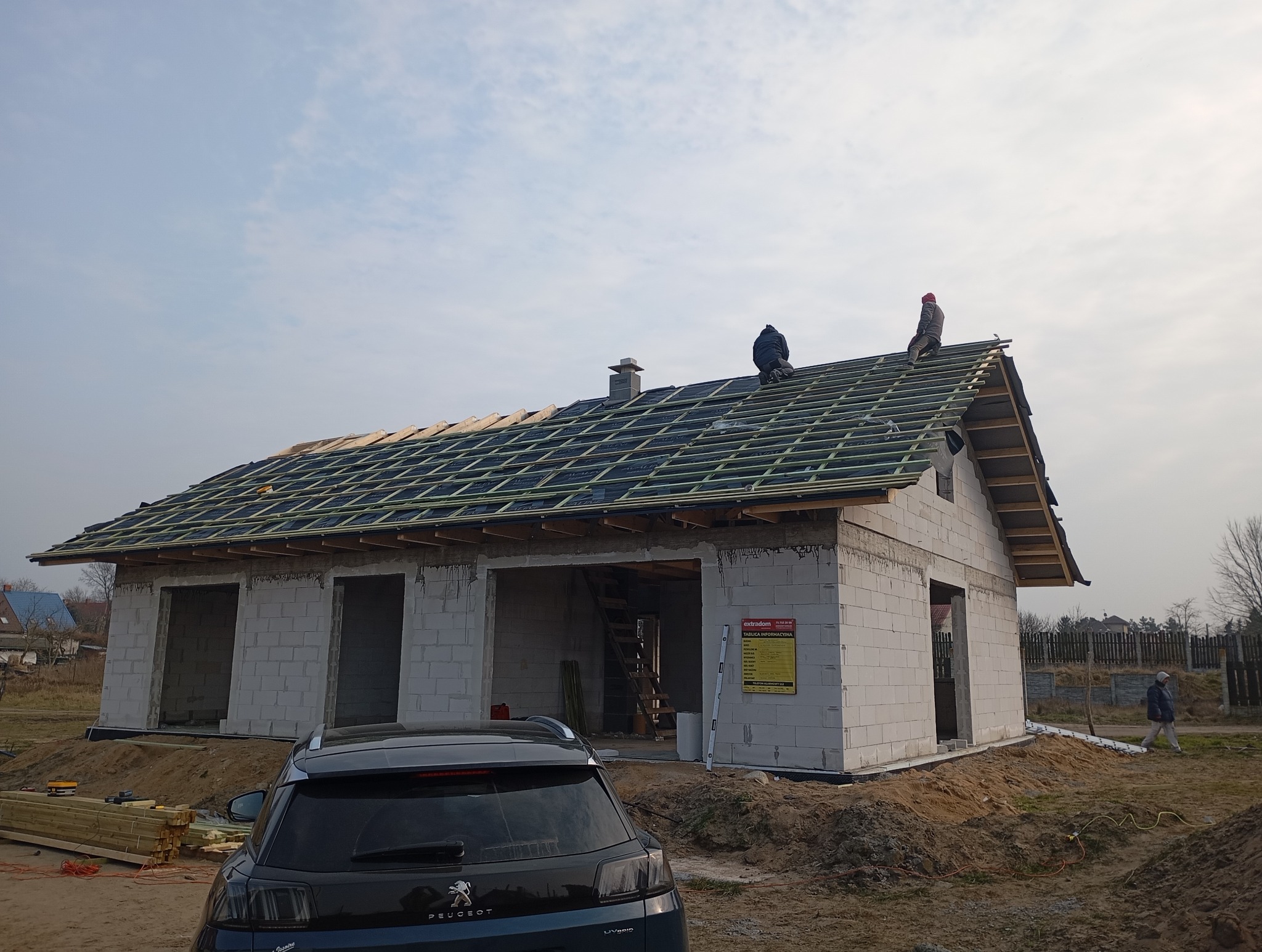 Budowa domu jednorodzinnego w stanie surowym otwartym: dekarze montują konstrukcję dachu z łat i kontrłat pokrytą membraną dachową, widoczny komin, ściany z białego bloczka, zaparkowany samochód...