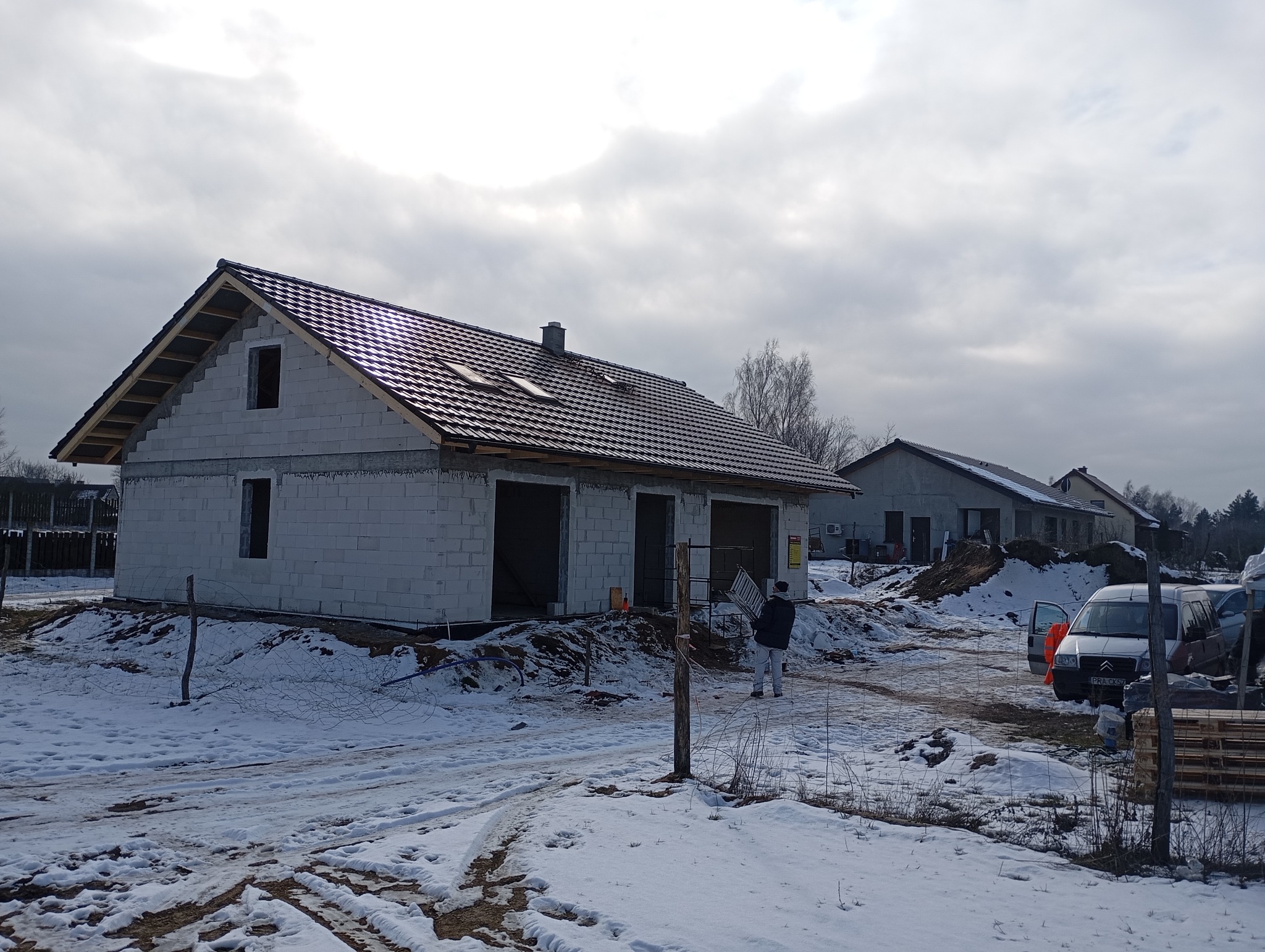 Dom w stanie surowym zamkniętym z zamontowanym dachem z dachówki ceramicznej, widoczne okna dachowe i elementy więźby, na pierwszym planie ośnieżony teren.