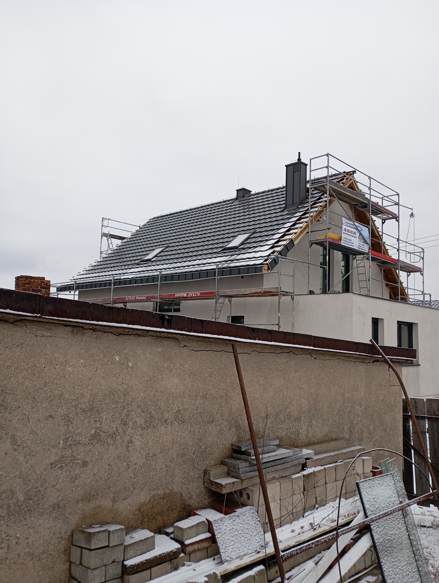Dom w trakcie krycia dachu z użyciem rusztowania. Widoczne dachówki, komin i okna dachowe. Częściowo ośnieżony dach i otoczenie. Na rusztowaniu baner reklamowy.