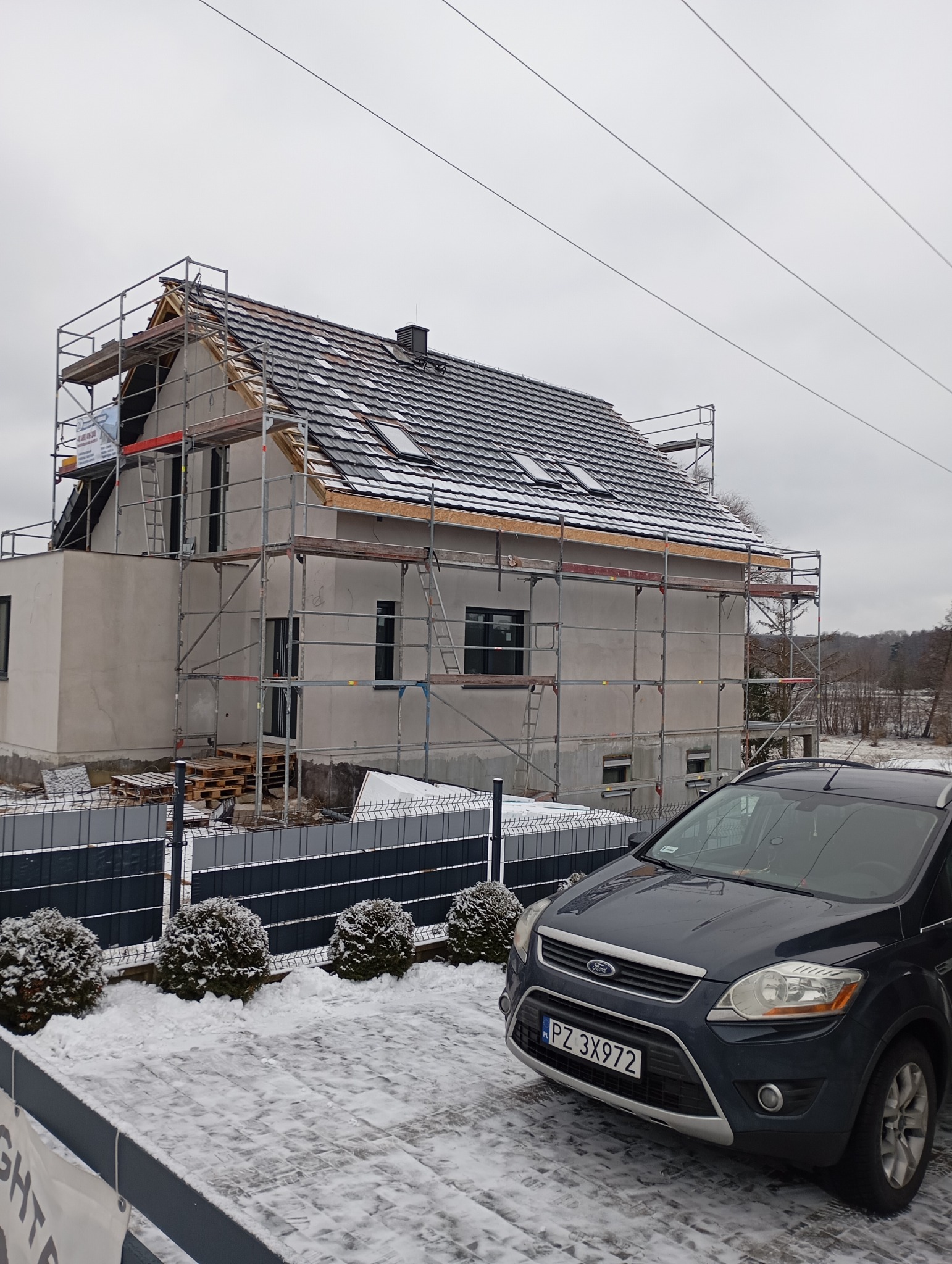 Dom w trakcie prac dekarskich, otoczony rusztowaniem. Częściowo pokryty dach z oknami dachowymi. Na pierwszym planie ośnieżony podjazd i samochód.