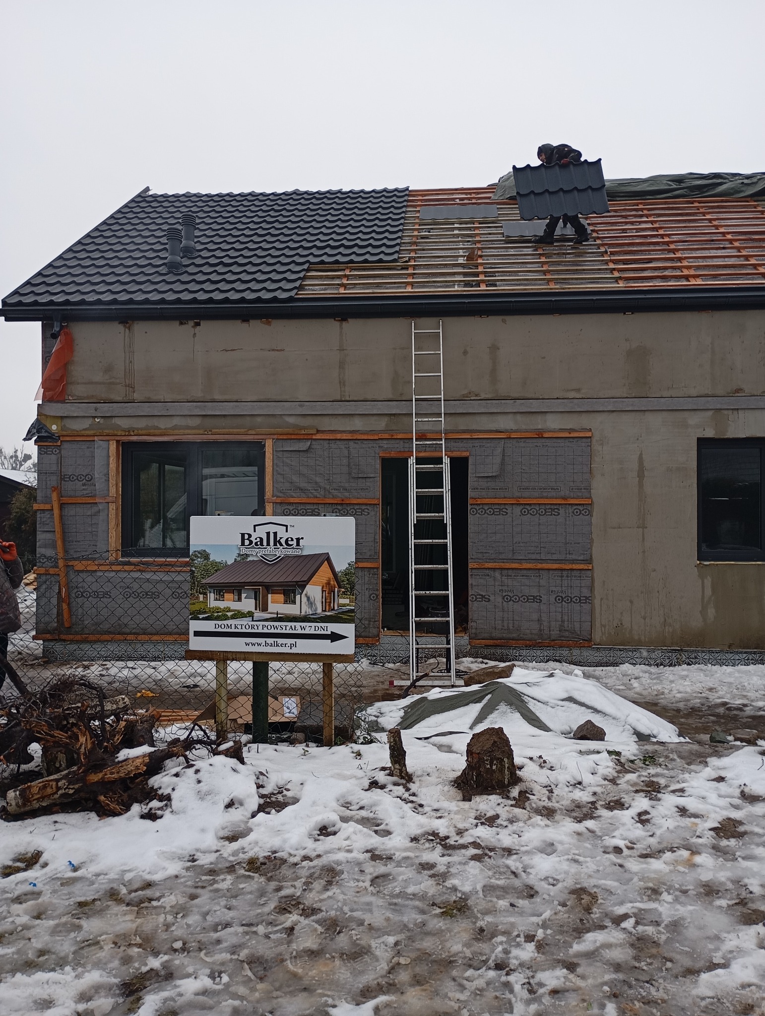 Układanie blachodachówki na dachu domu w Poznaniu. Widoczna drabina oparta o budynek i częściowo pokryty dach. Na pierwszym planie tablica informacyjna.