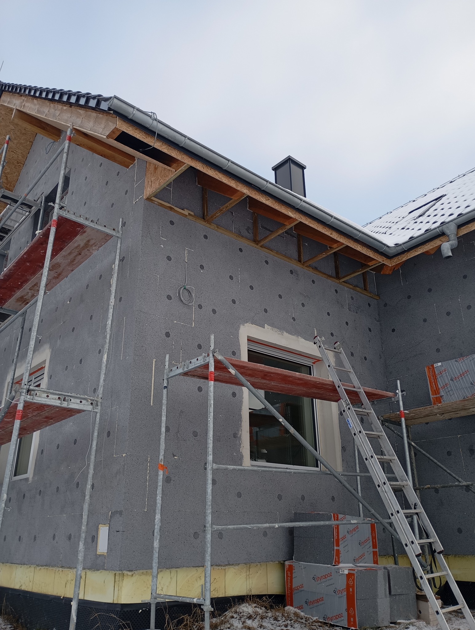 Dom w trakcie ocieplania: szare płyty styropianowe z charakterystycznymi kropkami, rusztowania, drabina oparta o ścianę, widoczne rynny i fragment dachu pokrytego śniegiem.