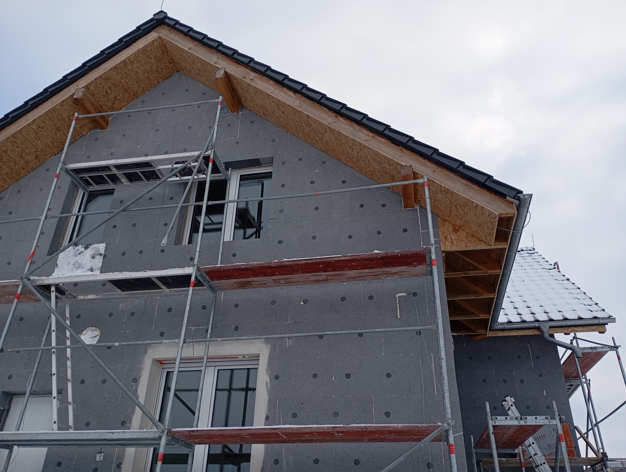 Dom w trakcie ocieplania, widoczne szare płyty izolacyjne na ścianach, rusztowania i fragment dachu z czarną dachówką. Część dachu pokryta śniegiem.