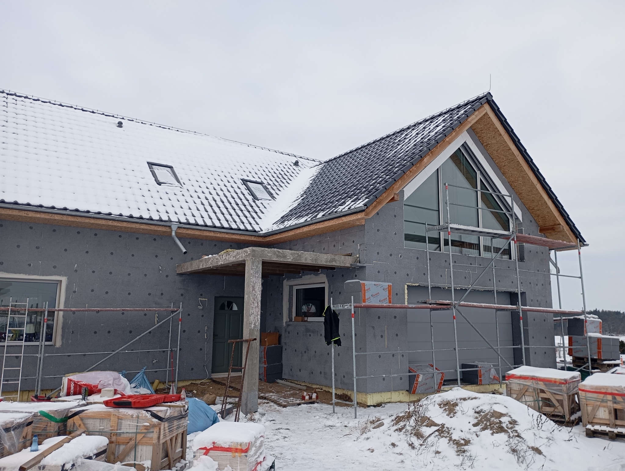 Dom w trakcie ocieplania styropianem grafitowym, częściowo pokryty dachem z dachówki ceramicznej. Widoczne rusztowania i materiały budowlane w zimowej scenerii.