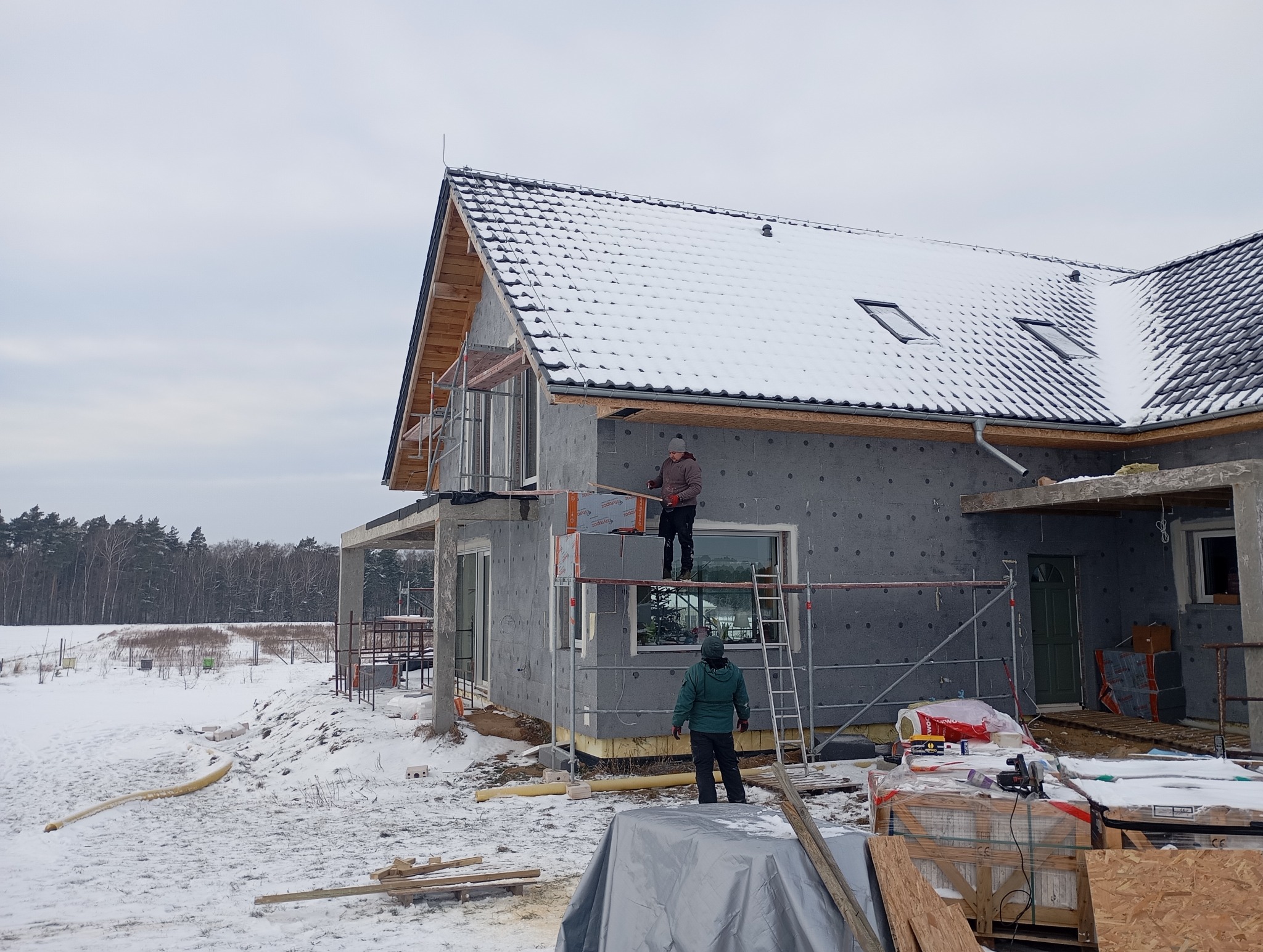 Dom w trakcie ocieplania, widoczne rusztowania i pracownicy montujący izolację. Dach pokryty śniegiem, zimowy krajobraz w tle.