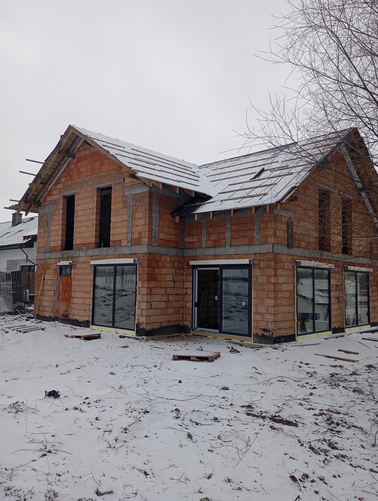 Dom w trakcie budowy, widoczne ściany z czerwonej cegły i częściowo wykonany dach pokryty śniegiem. Stolarka okienna zamontowana. Zima.