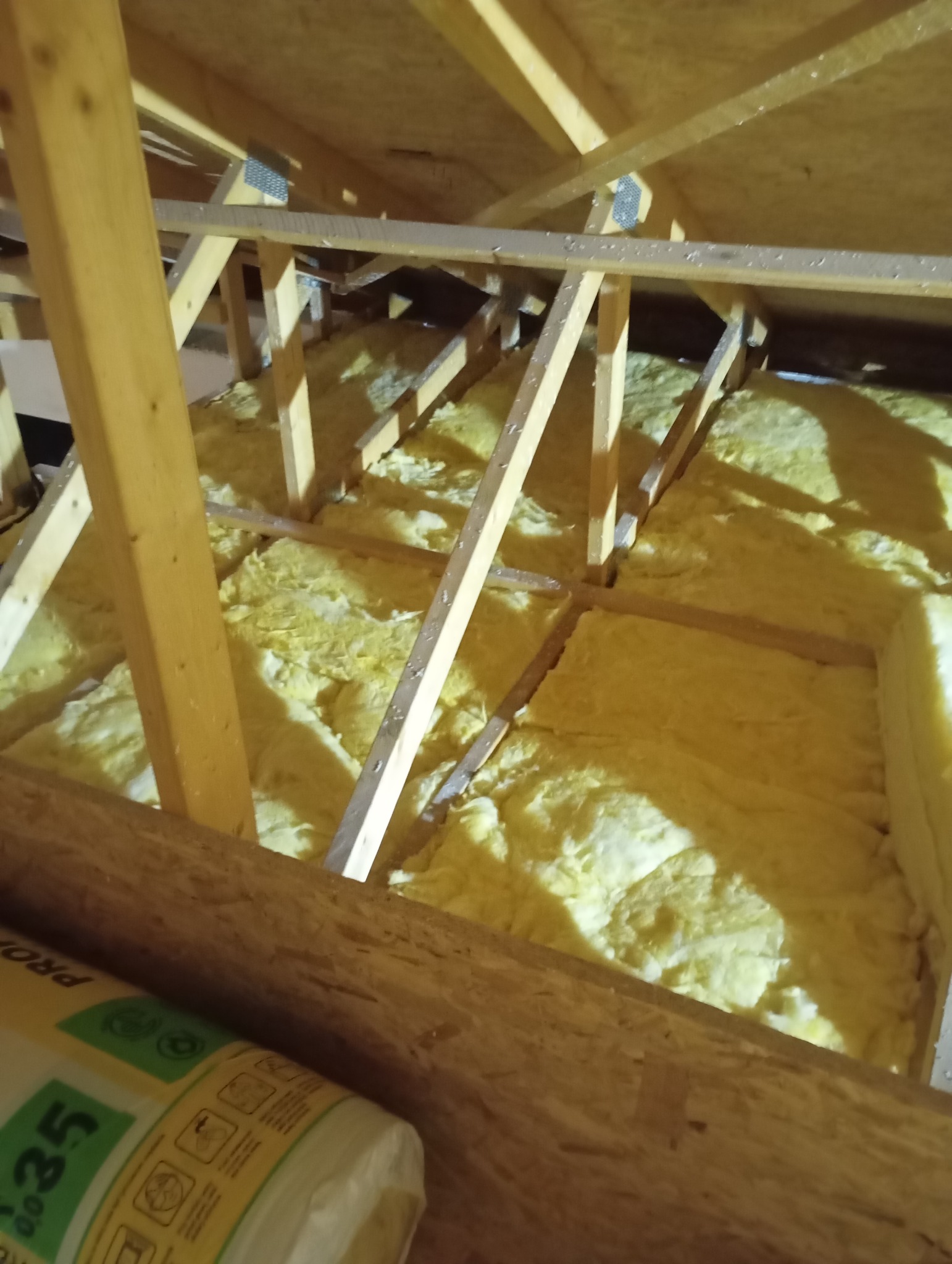 Żółta wełna mineralna między drewnianymi krokwiami poddasza, widok z dołu. Częściowo widoczna rolka izolacji. Oświetlenie ciepłe, sztuczne.