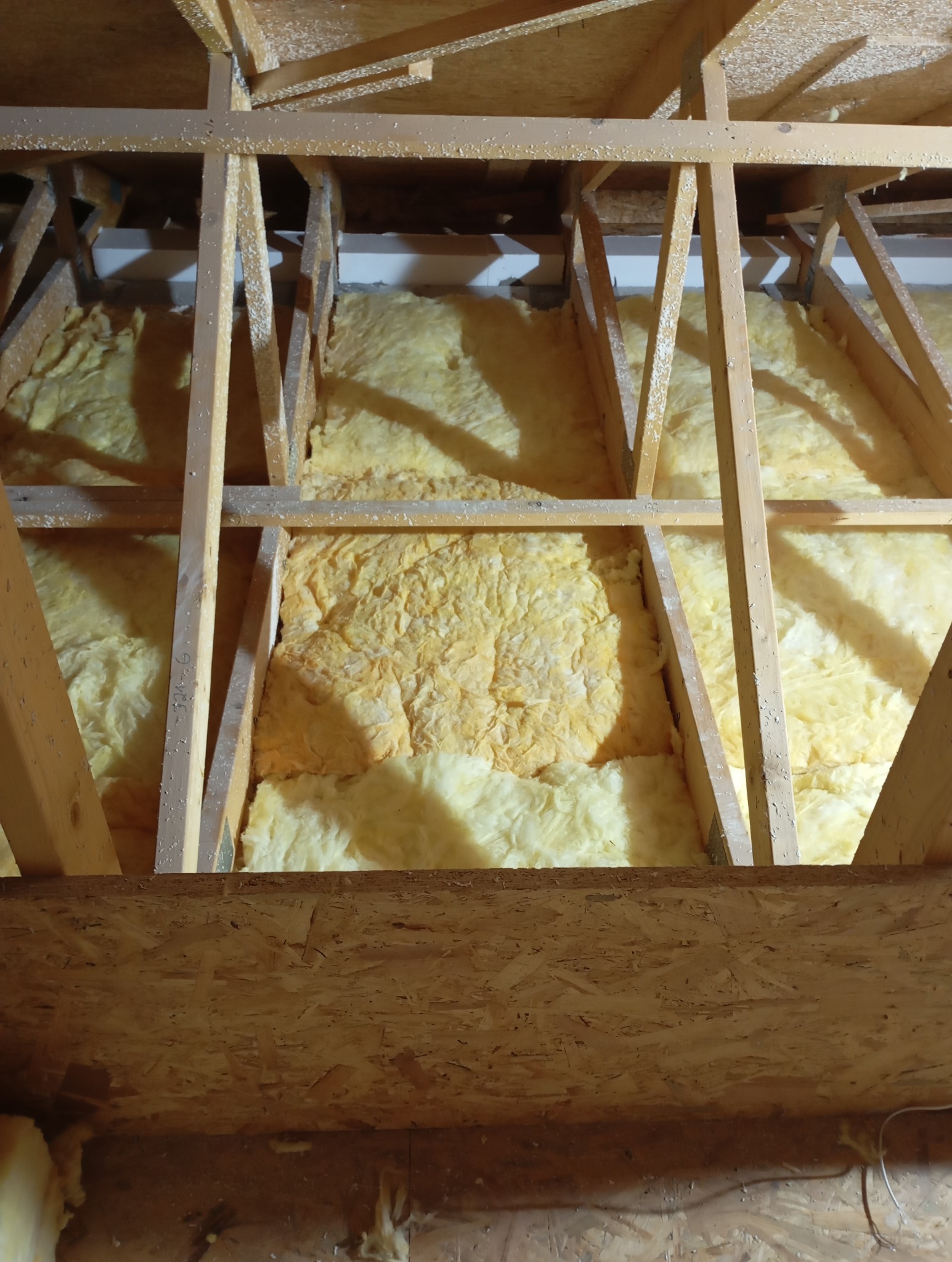 Ocieplenie poddasza wełną mineralną, widoczne drewniane elementy konstrukcyjne dachu. Fragment izolacji z bliska, ciepłe barwy oświetlenia.