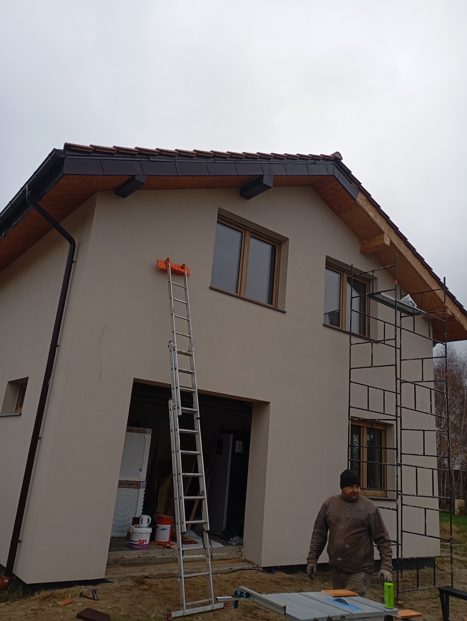 Dom jednorodzinny w trakcie prac wykończeniowych elewacji. Widoczne rusztowania, drabina oparta o ścianę, fragment dachu z brązową dachówką i pracownik w ubraniu roboczym.