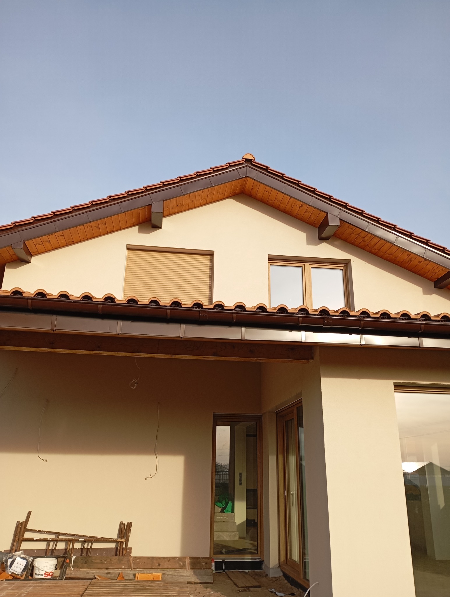 Dom jednorodzinny z brązową dachówką ceramiczną i drewnianą podbitką. Widoczne okna, jedno z roletą zewnętrzną. Częściowo widoczne rusztowanie i materiały budowlane.