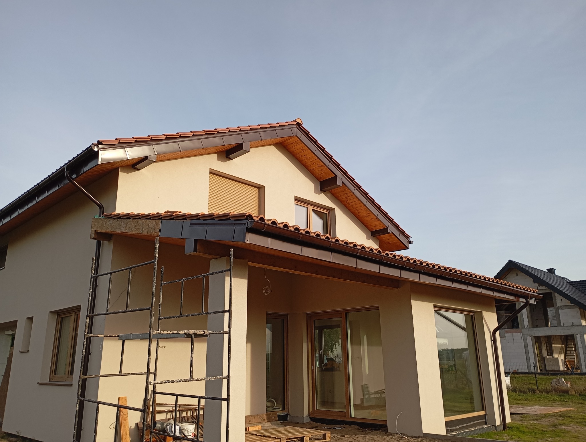 Dom jednorodzinny z nowym dachem z dachówki ceramicznej i zamontowanymi rynnami w kolorze brązowym. Widoczne rusztowanie przy elewacji.