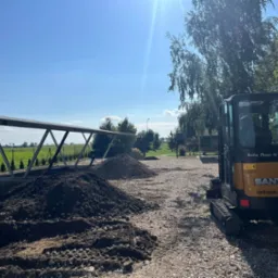 Żółto-czarna mini koparka SANY na żwirowym podłożu, obok pryzmy ziemi, w tle zadaszenie, zielona łąka i błękitne niebo z promieniami słońca.