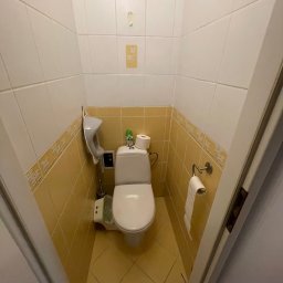 Sebastian Skwara - Wnętrze małej łazienki z toaletą, żółtymi płytkami i akcesoriami, widok zza uchylonych drzwi.