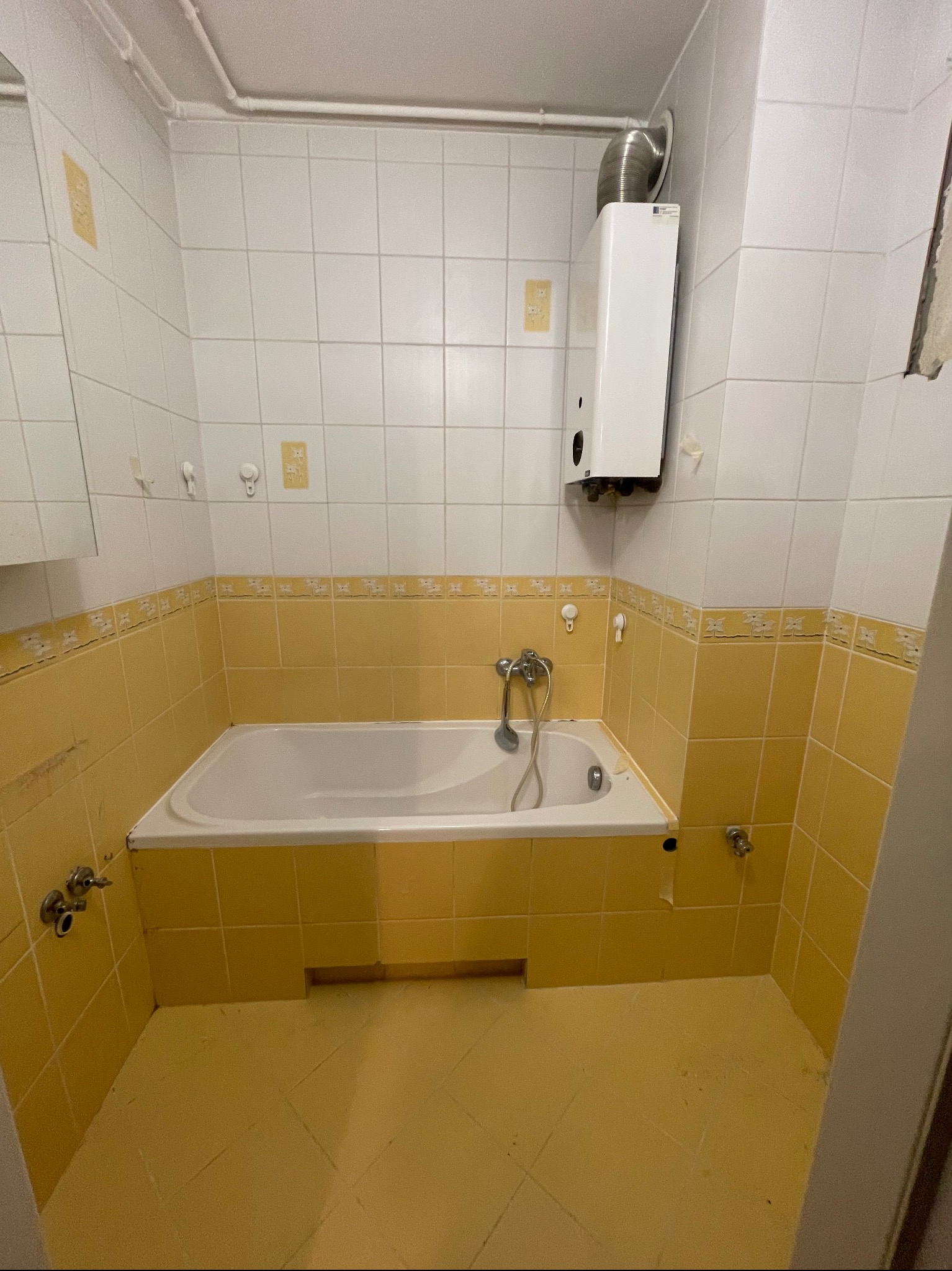 Żółto-biała łazienka z wanną, bojlerem i kafelkami z motywem kwiatowym. Widoczne ślady użytkowania i drobne uszkodzenia.