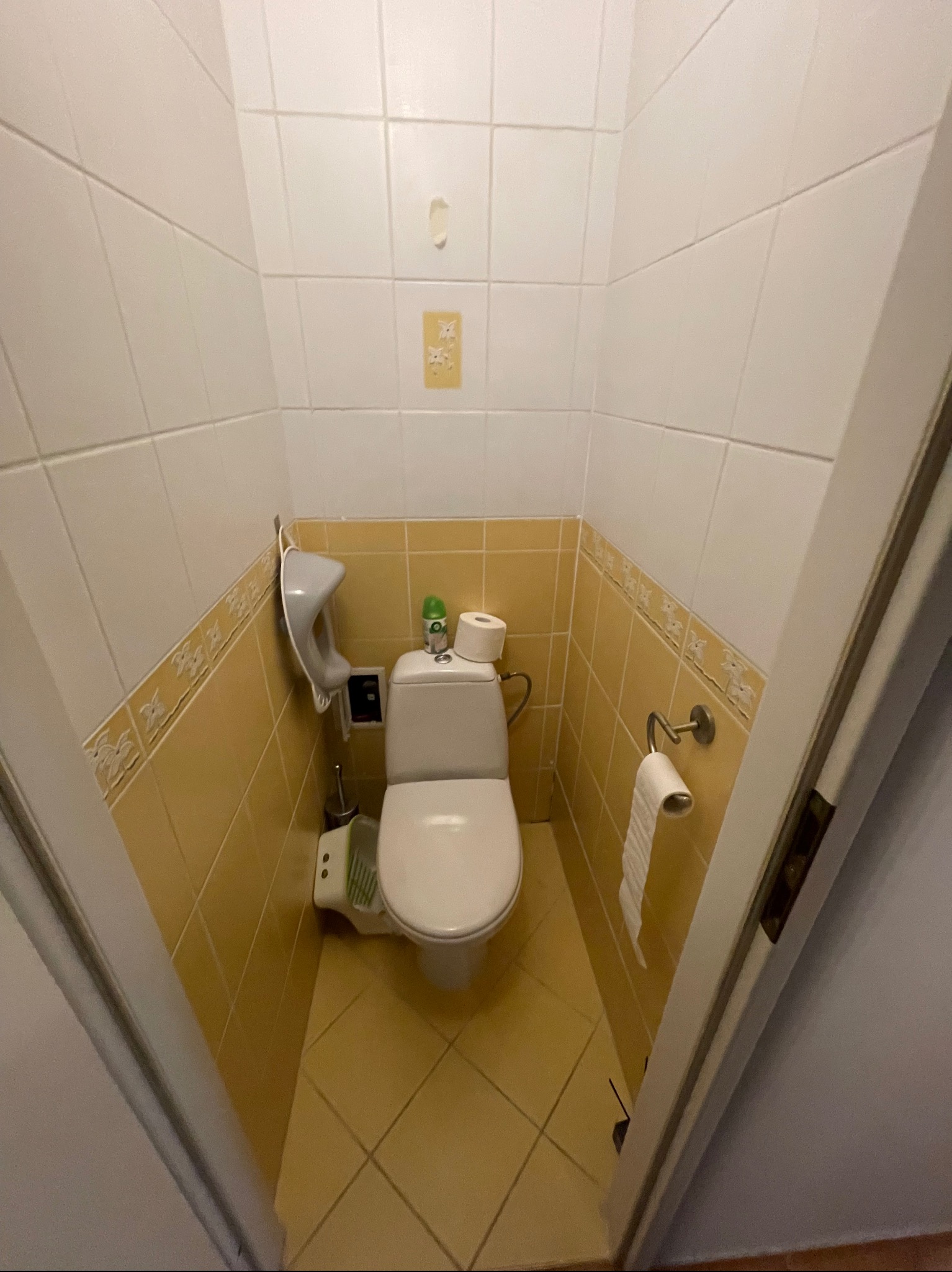 Wnętrze małej łazienki z toaletą, żółtymi płytkami i akcesoriami, widok zza uchylonych drzwi.