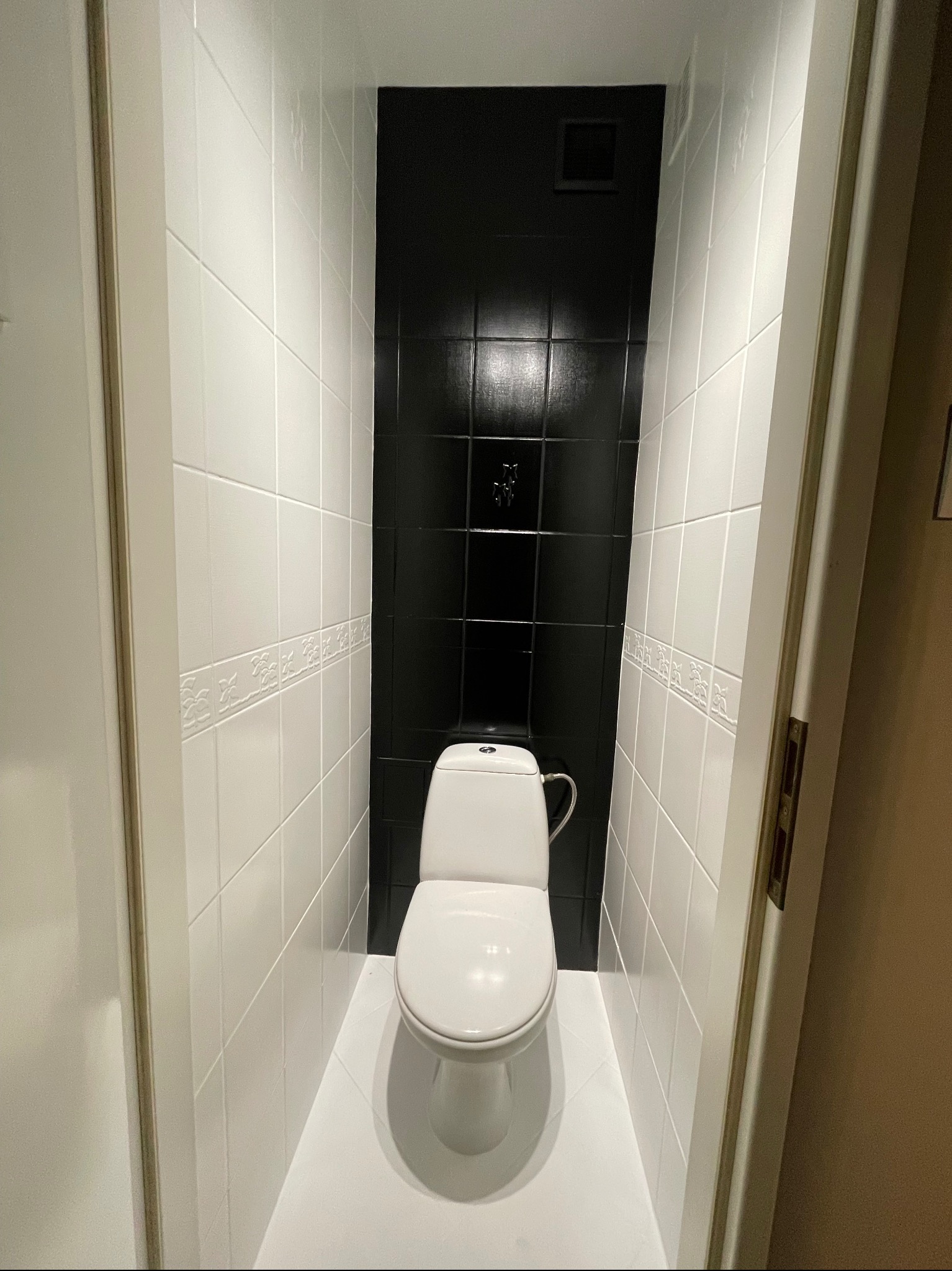 Wąska toaleta po remoncie: białe płytki z dekoracyjnym pasem, czarna ściana z kwadratowych płytek i biała podłoga.