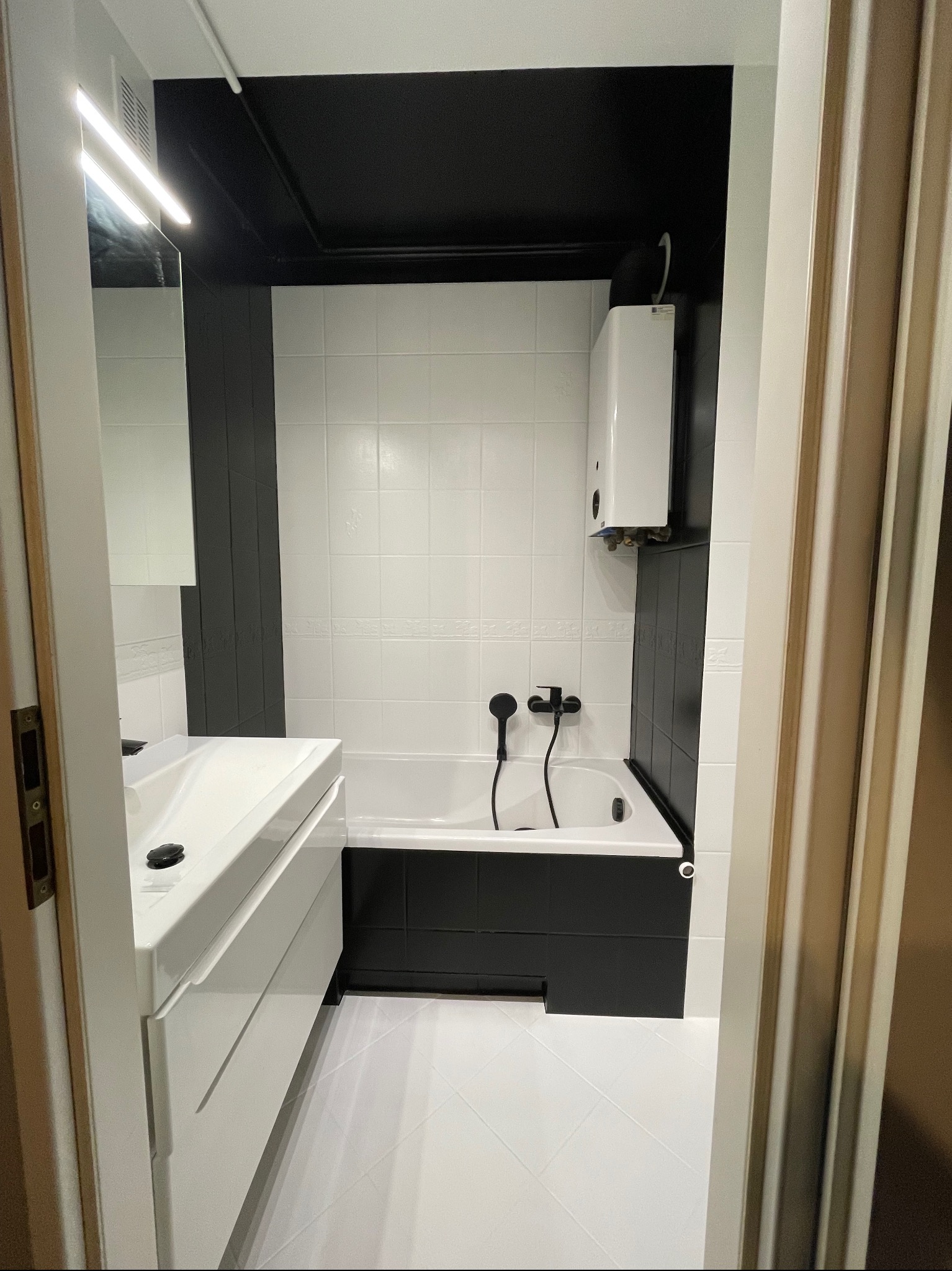 Wykończona łazienka w czarno-białej kolorystyce z wanną, umywalką i grzejnikiem wody. Czarny sufit i obudowa wanny kontrastują z białymi płytkami i podłogą.