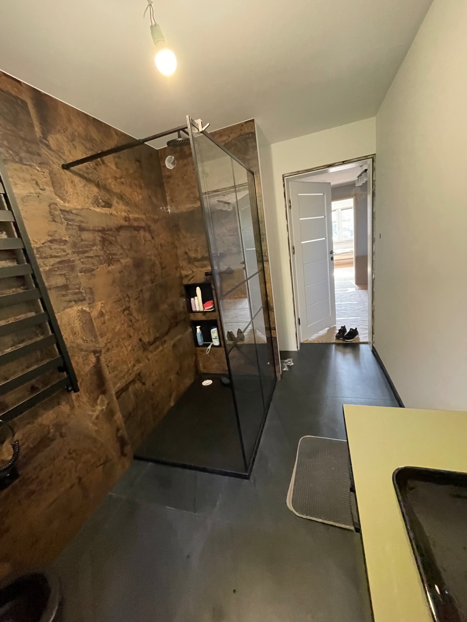 Wykończona łazienka z kabiną prysznicową typu walk-in, ściana wyłożona panelami imitującymi kamień, czarna armatura i podłoga, widoczna półka z kosmetykami wewnątrz kabiny, w tle otwarte drzwi...