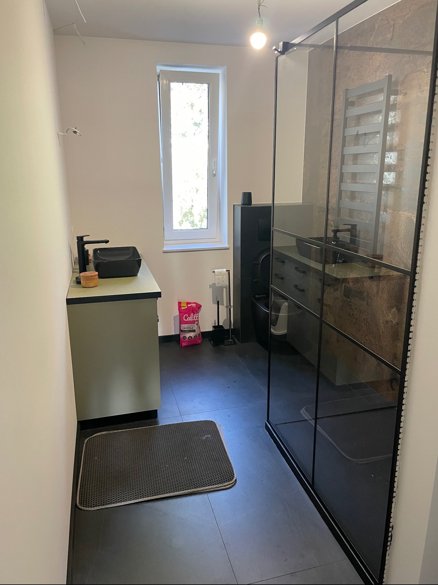 Nowoczesna łazienka z czarną armaturą, kabiną prysznicową typu walk-in z czarnymi szprosami, szarą podłogą, jasnymi ścianami i oknem. Na blacie umywalki stoi drewniany pojemnik, a obok toalety...