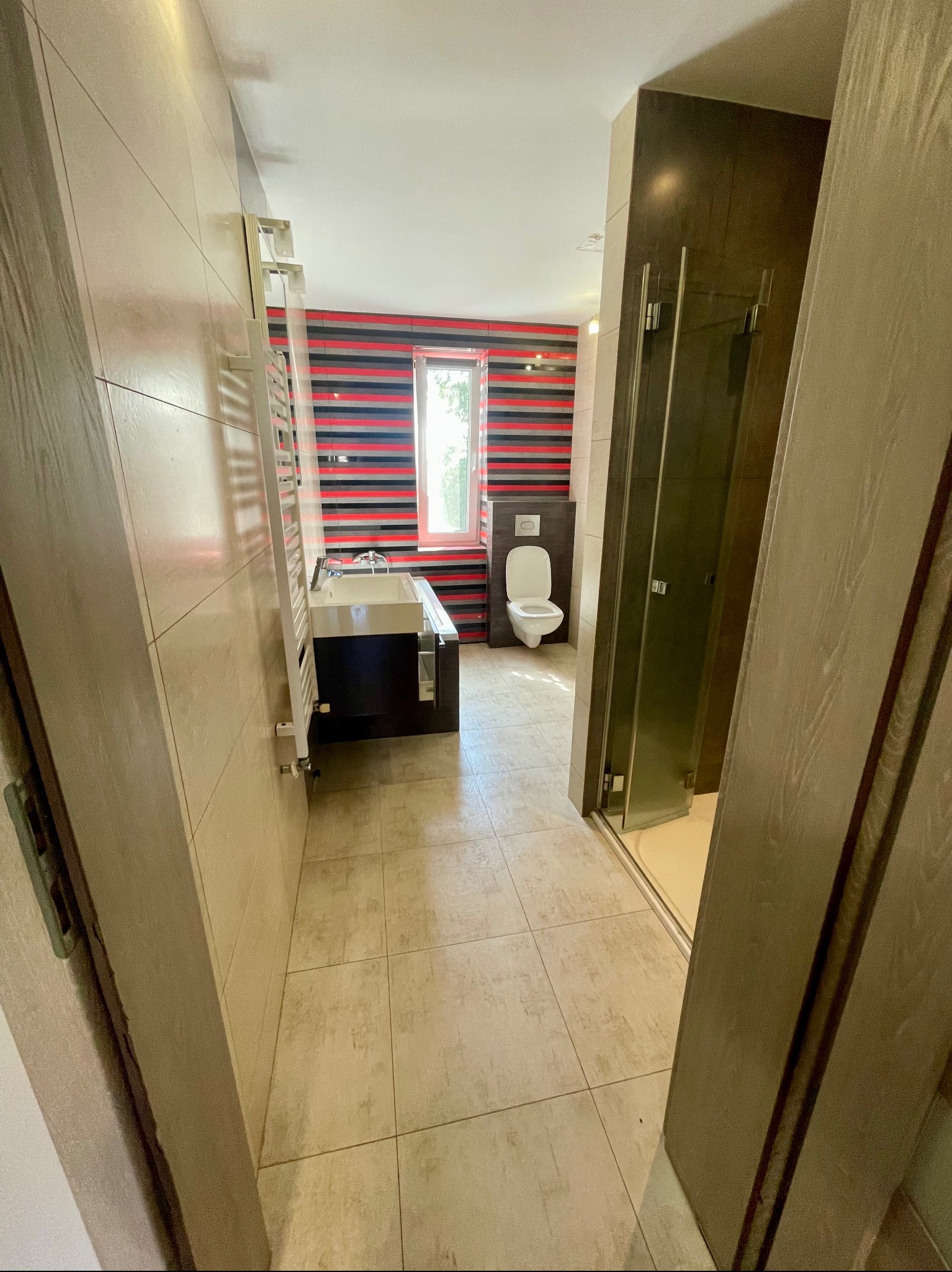 Ukończona łazienka z umywalką w zabudowie, toaletą wiszącą i kabiną prysznicową; ściana z poziomymi pasami czerwono-czarnych płytek za umywalką.