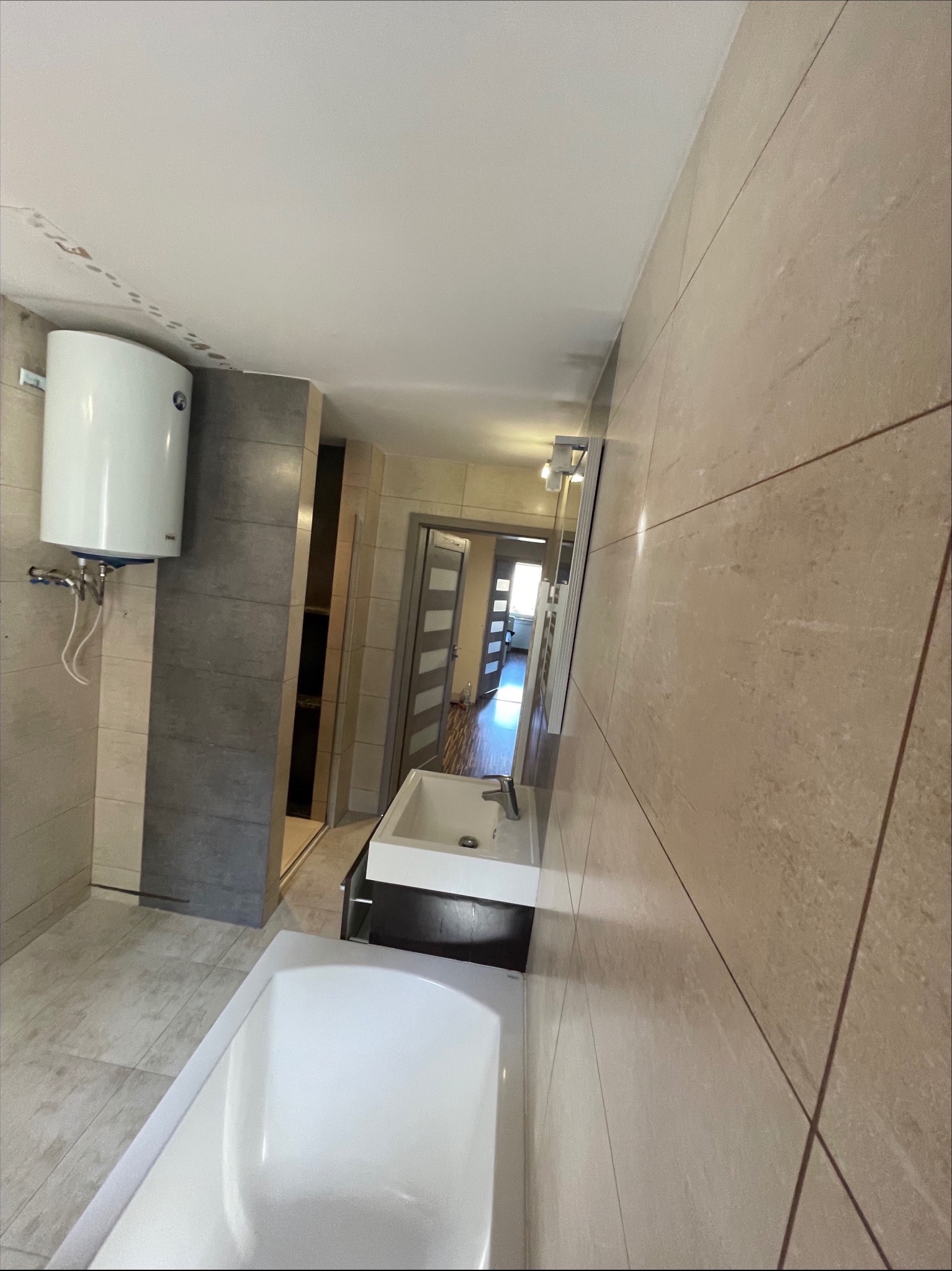 Nowoczesna łazienka z beżowymi płytkami imitującymi kamień, białą wanną, umywalką z szafką w kolorze ciemnego drewna i podgrzewaczem wody na ścianie.