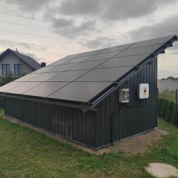 MDMS HOLDING - Twój partner od termomodernizacji (POZNAŃ) - Mały budynek gospodarczy z panelami fotowoltaicznymi na dachu, inwerter zamontowany na ścianie, w tle dom jednorodzinny i pochmurne niebo.