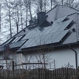 MDMS HOLDING - Twój partner od termomodernizacji (POZNAŃ) - Montaż paneli słonecznych na dachu domu z dachówką łuskową; dwaj monterzy na rusztowaniu, drabina oparta o ścianę, drzewa w tle.