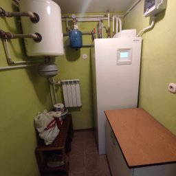 MDMS HOLDING - Twój partner od termomodernizacji (POZNAŃ) - Kotłownia z białą pompą ciepła Hitachi, zbiornikiem wyrównawczym, grzejnikiem i szafką, widok na ścianę w odcieniu zieleni.