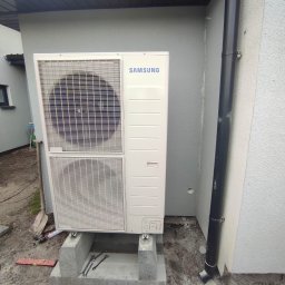 MDMS HOLDING - Twój partner od termomodernizacji (POZNAŃ) - Biała pompa ciepła Samsung zamontowana na betonowej podstawie przy ścianie budynku z rurą spustową, widoczne narzędzia i poziomica sugerujące trwający montaż.