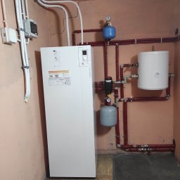 MDMS HOLDING - Twój partner od termomodernizacji (POZNAŃ) - Biała pompa ciepła Atlantic w piwnicy z rurami w kolorze cegły, zbiornikiem wyrównawczym i manometrem.