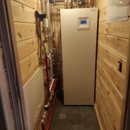 Wnętrze kotłowni z drewnianą boazerią, widoczna biała pompa ciepła Hitachi z niebieskim wyświetlaczem, liczne rury i zawory, oraz niebieski zbiornik wyrównawczy.