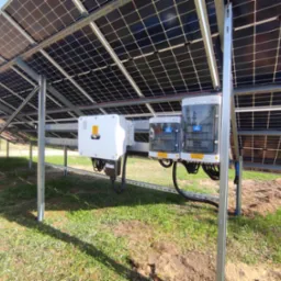 Widok od dołu na instalację fotowoltaiczną z widocznymi panelami, inwerterem i skrzynkami zabezpieczeń na tle trawy.