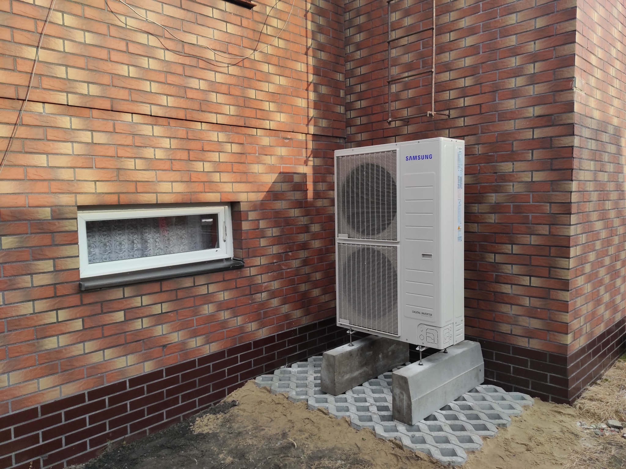 Biała jednostka zewnętrzna pompy ciepła Samsung Digital Inverter zamontowana na betonowych blokach przy ceglanym budynku z małym oknem.