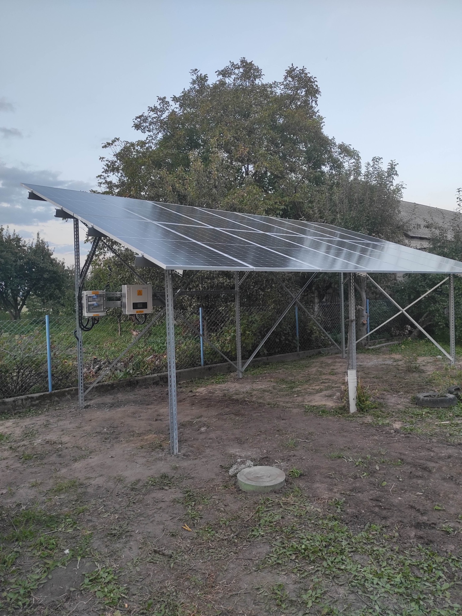 Panel fotowoltaiczny na metalowej konstrukcji w ogrodzie, widoczne skrzynki elektryczne, w tle drzewa i ogrodzenie z siatki.
