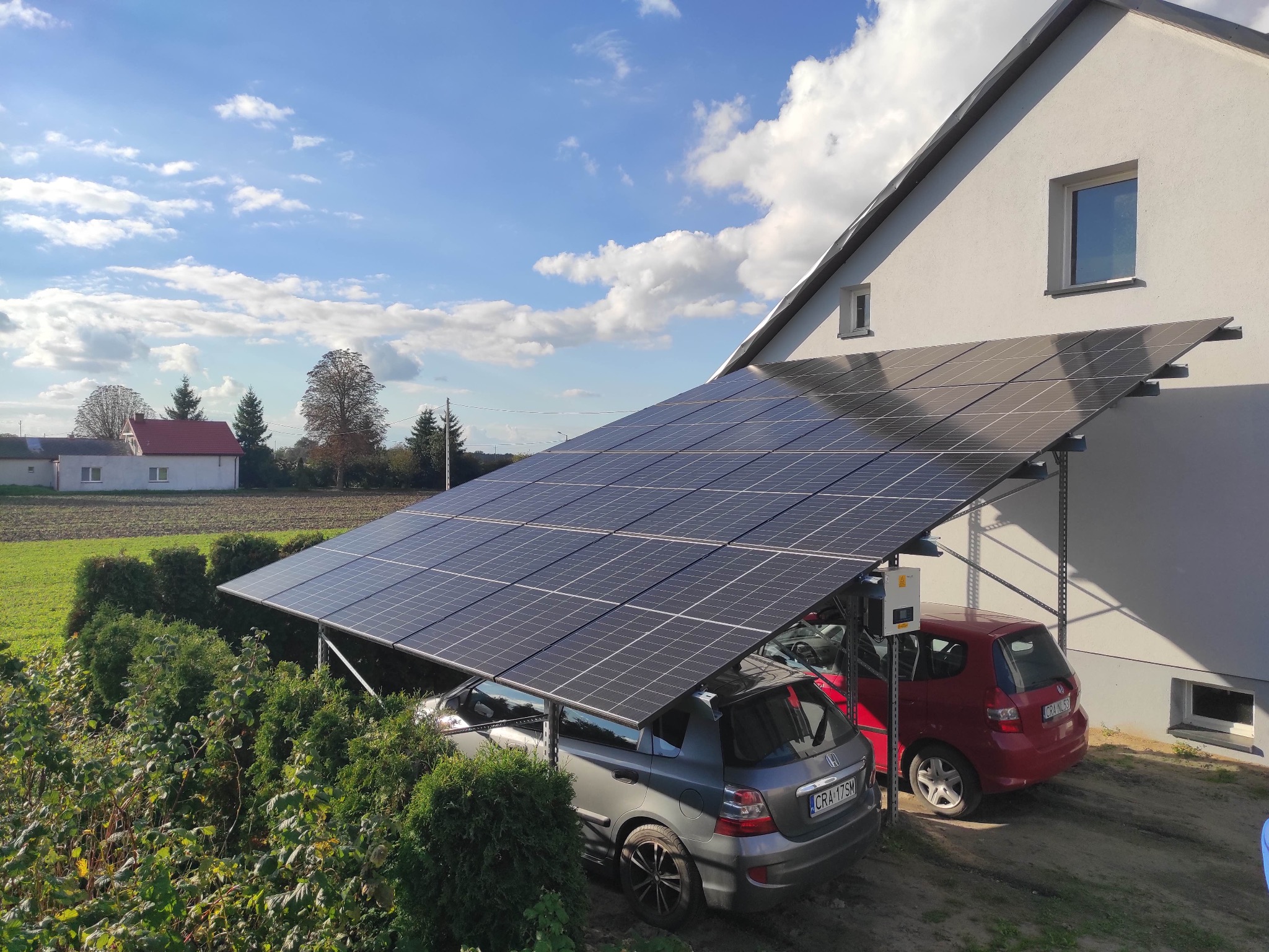 Instalacja fotowoltaiczna jako wiata garażowa dla dwóch samochodów, z widoczną skrzynką elektryczną na ścianie budynku i zielonym otoczeniem.